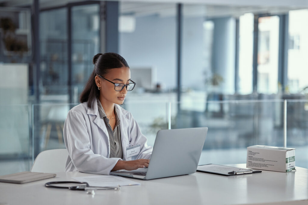 A female doctor in a white lab coat works at her laptop in a glass-walled conference room.